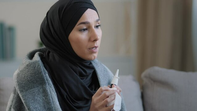 Muslim arabian woman sick islamic girl in hijab sit on cozy sofa at home treating runny nose use medicine nasal spray cure snuffle daily therapy from seasonal allergy symptoms recover sickness allergy