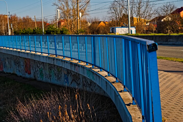 Poręcz stalowa , mostu drogowego i pieszego pomalowana na niebiesko mi wygięta w łuk . Betonowa ściana pokryta graffiti .
