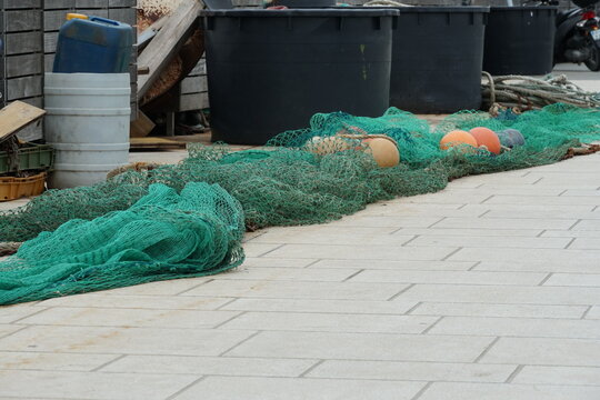 Green fishing net with red-brown floats stretched on the tiled pavement in port ready for loading on fishing boat. Behind the net are black and white plastic buckets. 