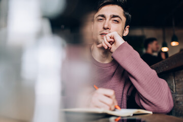 Portrait of intelligent male student with education textbook looking at camera while learning in coworking space, smart hipster guy with copybook for doing course work or university test posing