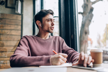 Contemplative Caucaisan student in electronic earbuds listening audio book and thinking about education learning in coffee shop, millennial hipster guy in bluetooth headphones creating content plan