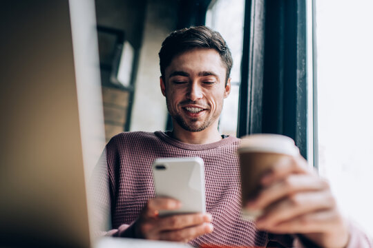 Cheerful IT professional with takeaway caffeine beverage and smartphone gadget watching funny video, happy Caucasian hipster guy connecting to bluetooth for sharing media via mobile app and laptop