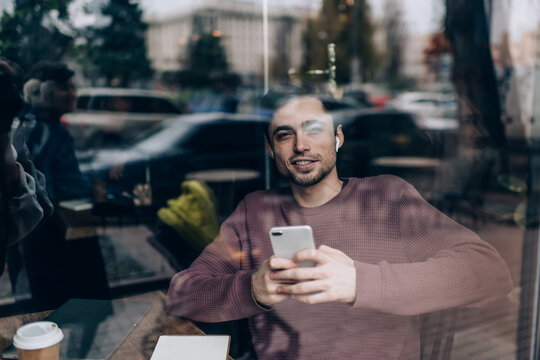 Portrait of Caucaisan male blogger in headphones using media application on cellphone gadget, happy hipster guy in bluetooth earbuds listening playlist during leisure pastime in cafe interior
