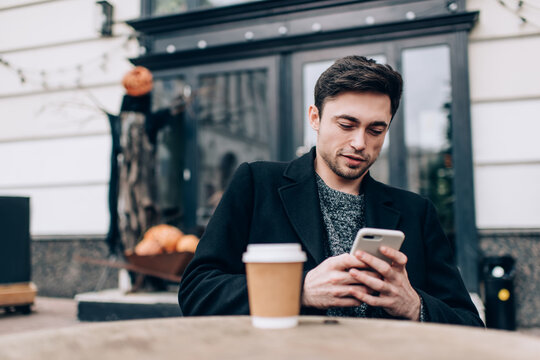 Millennial travel blogger with takeaway caffeine beverage browsing wireless website on modern cellphone technology, carefree hipster guy making online booking via mobile application using 4g internet