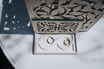 A white wooden box with a pattern, a symbol of infinity with gold rings, lies on a white table at a wedding ceremony. Festive photo, close-up.
