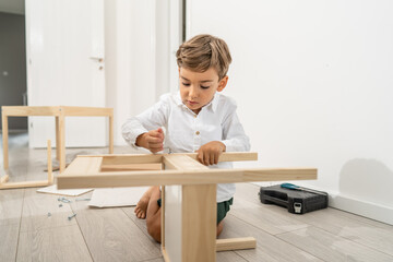 Naklejka premium Small caucasian boy four years old playing with self assembly chair or table on the floor at home in day - little child assembling furniture alone childhood craft concept front view copy space