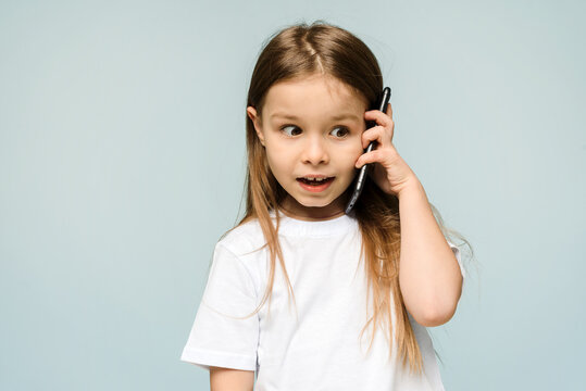 OMG, shocked little schoolgirl holding a smartphone to her ear on a blue background in the studio. A little girl is talking on the phone and is surprised. copy space