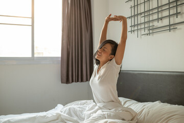 Asian woman wake up in the morning, sitting on white bed and stretching, feeling happy and fresh. happy millennial girl lying on soft pillows awaken in cozy bedroom, relaxation concept