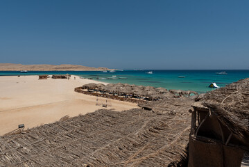 Holidays in Egypt. Beach with umbrellas made of dry palm leaves, desert and yachts in the blue water of the Red Sea. Vacation and Holidays in Egypt.