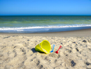 Fototapeta na wymiar A yellow sand bucket and colorful toy shovels at the beach by the ocean.