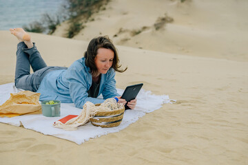  young woman on the beach by the ocean in spring sits on a picnic, reads a book on a tablet, watches a movie, podcast, modern technologies