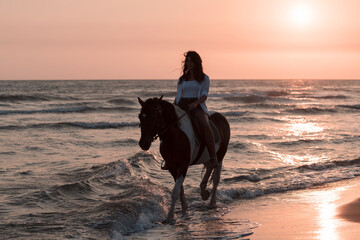 Woman in summer clothes enjoys riding a horse on a beautiful sandy beach at sunset. Selective focus  - obrazy, fototapety, plakaty