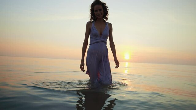 Full body view: Beautiful WET woman in see through dress walking at you against sunset sea background