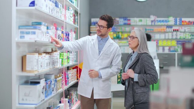 Caucasian male pharmacist in white lab coat and glasses helping female customer in modern drugstore. Senior woman consulting with professional apothecary about medicine dosage.