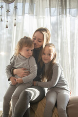 Portrait of a beautiful young woman with two little daughters at home against the background of the window. Family spend leisure time in cozy home environment