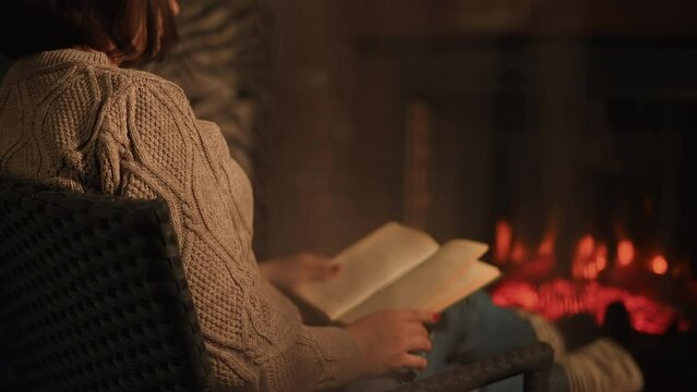 A woman sits by the fireplace in the evening and reads a book