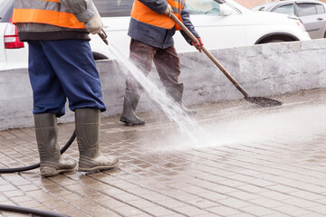 Two Man, workers in yellow orange vest and rubber boots washing sidewalk, road. Cleaning street...