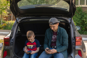 father and son ignore each other using their smartphones. they sit in the trunk of the car and do not pay attention to each other. concept of modern smartphone addiction