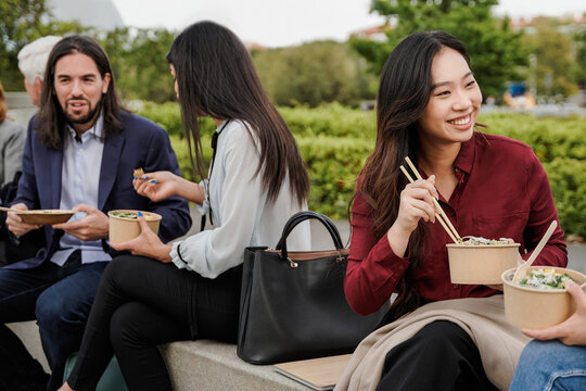 Multiethnic business people eating takeaway food during lunch break outdoor outside the office - Focus on Asian woman face