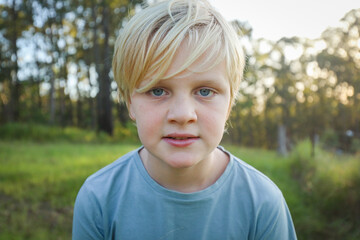 Blonde boy with grumpy expression close up in the bush at sunset