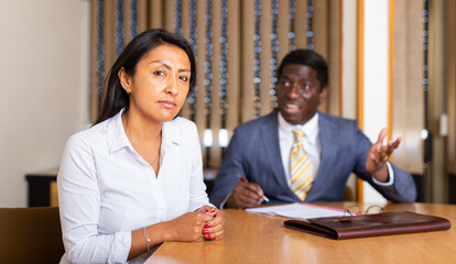 Frustrated hispanic woman entrepreneur sitting at office desk on background with angry partner...