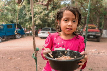 Latin girl sitting on a swing eating from a plastic bowl. Concept of childhood, nutrition and food in rural areas - obrazy, fototapety, plakaty