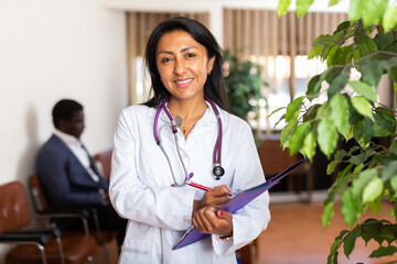 Smiling hispanic female general practitioner in white uniform standing in clinic, filling clipboard...