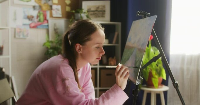Young woman artist close-up, sitting and painting beautiful picture landscape on easel using watercolour and brush in workshop. Female painter drawing illustration with aquarelle paint in art school. 