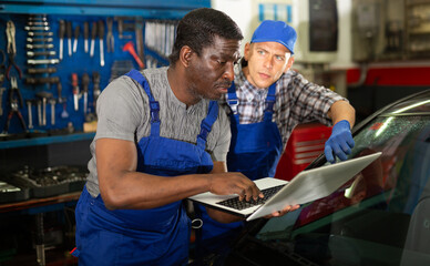 Mechanic man manager with worker in a workshop checks the electronics of the car using laptop