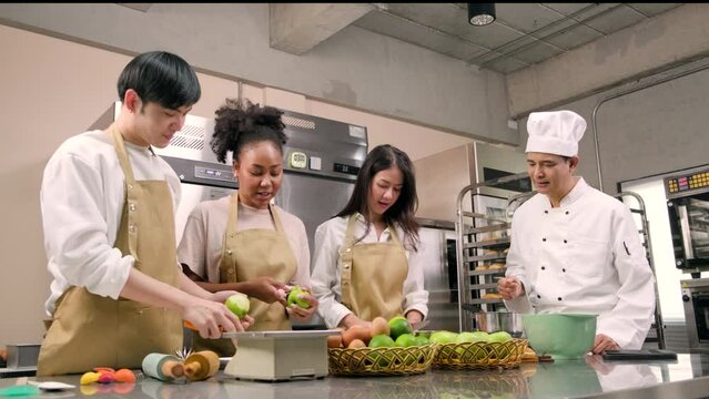 Hobby cuisine course, senior male chef in cook uniform teaches young cooking class students to peel and chop apples, ingredients for pastry foods, fruit pies in restaurant stainless steel kitchen.