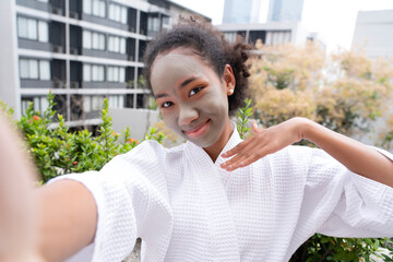Young African American woman shooting a selfie with smartphone after applying mud mask with smiling face to post her activities on social media, self-portrait action charming in front of camera.