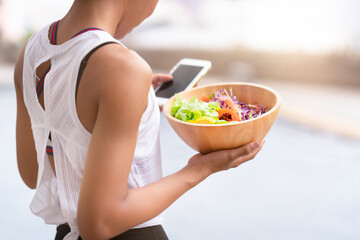 Young black woman touch mobile screen and holding salad bowl in hands after workout at home, chatting with friends, check incoming mails, searching online shopping website to buy sport equipment