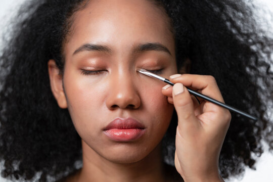 Makeup artist applies eye shadow on eyelid of African American model by using makeup brush to spread the color.