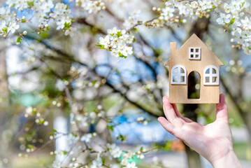 The girl holds the house symbol against the background of blossoming cherry

