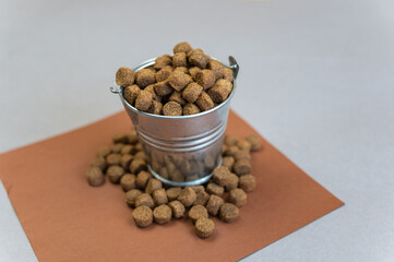 A full metal bucket of dry pelleted dog food. A small silver thigh of dog food stands against a gray background. A square brown piece of paper with a scattering of pellets.