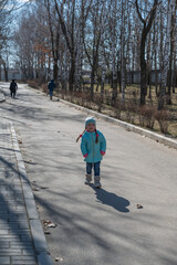 Fototapeta na wymiar A child having fun in a sunny spring park. A five-year-old girl with long pigtails in a turquoise jacket poses and curses. A birch tree without leaves in the background