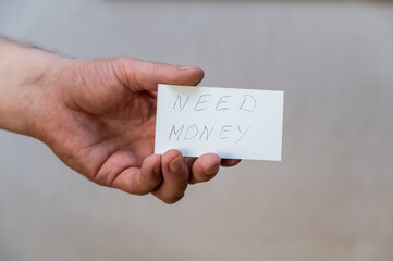 Need money. Words written in jagged letters. A man's hand holds a white paper rectangle with text against a gray background.