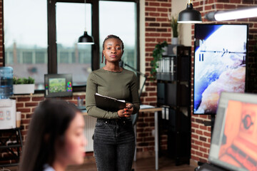 Production department african american team leader with clipboard standing in modern interior. Creative agency office confident worker standing in workspace, looking at camera.