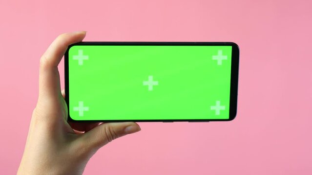 close-up of female hands holding smartphone with blank green screen showing copy space for app or website design, on pink background , mockup.