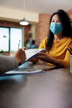 Businesswoman with medical face mask holding clipboard while freelancer signing papers for executive manager job during company interview. Team working at business collboration in startup office