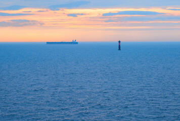 Obraz premium seascape in the evening in cloudy weather after sunset in the blue hour, a lot of water and a ship in the haze sailing past the lighthouse, the clouds illuminated by the gold of the sun's rays