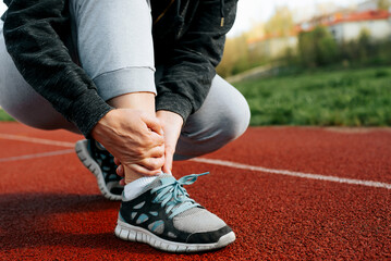 Woman runner holding leg suffering from muscle and tendon sprain pain in ankle at outdoor stadium, close-up. Athlete injured during intense workout. Health care and sport concept - obrazy, fototapety, plakaty