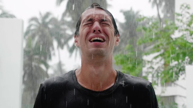 Portrait of crying sad man standing in the pouring rain and getting soaked. Slow motion shot
