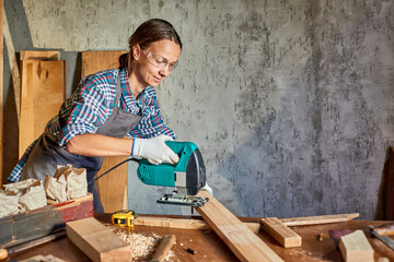 Female Carpenter sawing wood with an electric jigsaw. Woman in apron doing some carpentry work in his workshop or garage, using electric jigsaw.