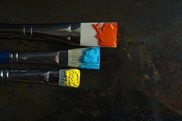Top down view of three horizontal paint brushes with primary yellow, teal and phthalo green oil paint on bristles. Dark grey grunge background with copy space.