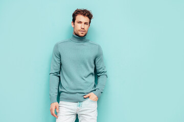 Fototapeta premium Portrait of handsome confident model. Sexy stylish man dressed in sweater and jeans. Fashion hipster male with curly hairstyle posing near blue wall in studio. Isolated