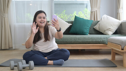 Cheerful overweight woman sitting on mat in living room and making video call via her smart phone.