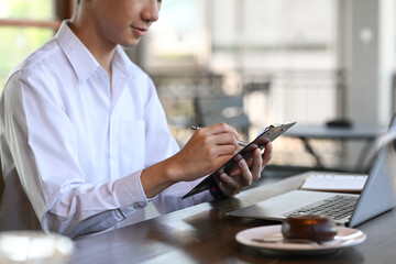 Young male office worker sitting in front of computer laptop and making notes on notepad.