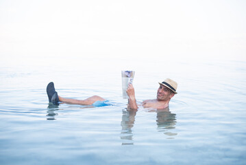 Man lies in blue water of Dead Sea. Person in hat and with newspaper. Very salty water push out....