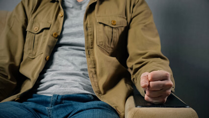 Man gets angry hitting armrest with fist sitting in soft armchair against grey wall at home. Male person shows expression of irritation close view